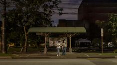 two people standing on the sidewalk in front of a building at night with street lights