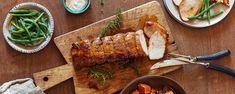 a wooden cutting board topped with meat and veggies next to plates of food