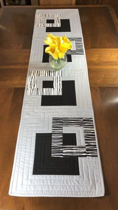 a table runner with yellow flowers in a glass vase on top of it and black and white designs