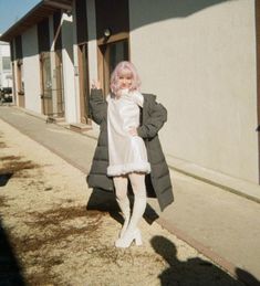 a woman with pink hair standing in front of a building wearing a white dress and coat