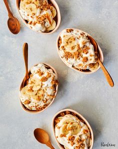 three bowls filled with bananas and granola on top of a gray surface next to wooden spoons