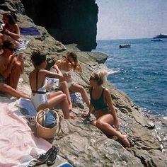 several people sitting on rocks near the ocean with boats in the water and one person wearing a bathing suit