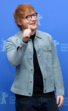 a man with red hair and glasses pointing at the camera while standing in front of a blue wall