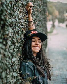 a woman wearing a hat leaning against a wall covered in vines and ivys smiling at the camera