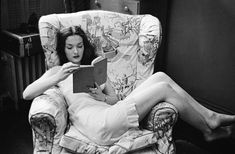 a woman reading a book while sitting in a chair with her feet up on the arm rest