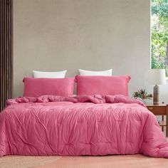 a bed with pink comforter and pillows in a room next to a window,