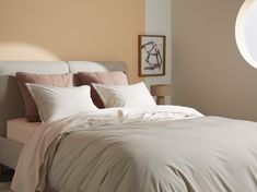 a bed with white sheets and pillows next to a round window in a beige room