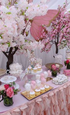 a table topped with cakes and cupcakes next to pink trees filled with flowers