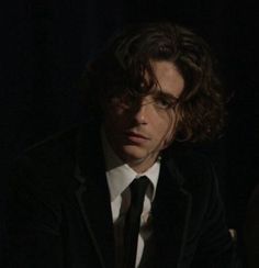 a man with long hair wearing a suit and tie sitting in front of a dark background