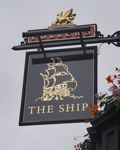 a sign that says the ship hanging off the side of a building with flowers in front of it