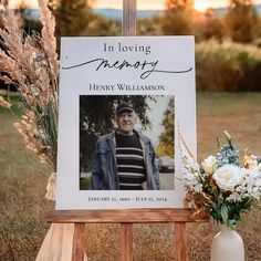 a memorial sign with flowers in front of it and a photo on the easel