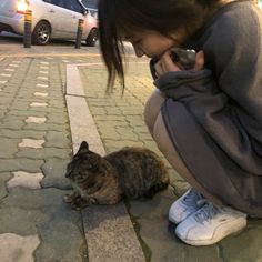 a woman kneeling down petting a cat on the sidewalk next to a parked car