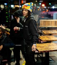 a young man standing in front of a wooden table wearing a beanie and holding a drink