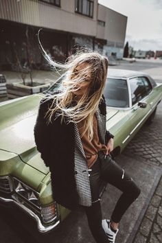 a woman sitting on the hood of a green car in front of a building with her hair blowing in the wind