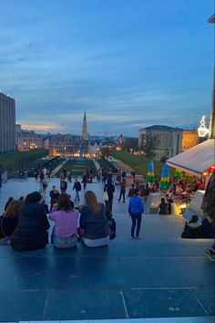 several people sitting on the ground in front of some buildings at dusk, one person is taking a photo