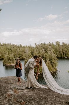 a bride and groom kissing on the edge of a cliff overlooking a lake with trees in the background