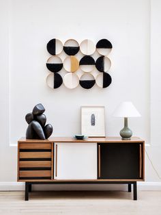 a wooden table topped with a lamp next to a wall mounted art piece on top of a white wall