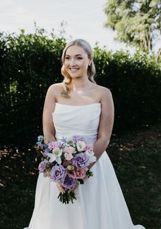 a woman in a white dress is holding a purple and pink bouquet on her wedding day