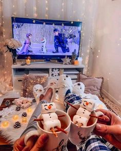 two people are holding mugs with marshmallow faces on them in front of a television