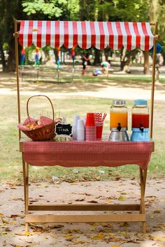 a picnic table with drinks on it in the middle of a park, next to a red and white striped awning