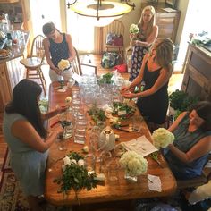 a group of women standing around a wooden table with wine glasses and flowers on it