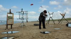 a woman standing on top of a sandy beach next to an easel and paintings