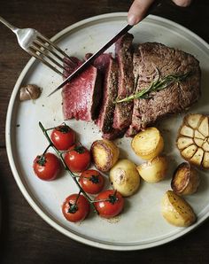 a person cutting up some meat on top of a plate with potatoes and cherry tomatoes