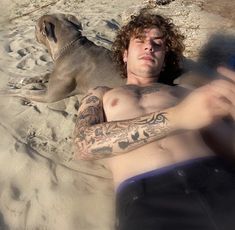 a man laying in the sand next to a dog with tattoos on his arm and chest