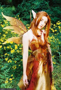 a woman dressed as a fairy standing in front of yellow flowers and trees with her hands on her hips