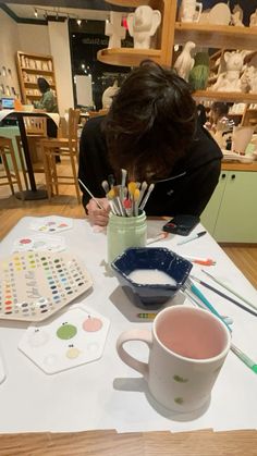 a woman sitting at a table working on some art work with paint and pencils