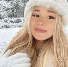 a woman with long blonde hair wearing a white hat and smiling at the camera in front of snow covered trees