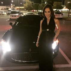 a woman standing next to a black car in a parking lot at night with headlights on