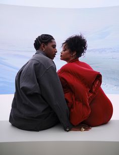 a man and woman sitting next to each other on top of a white wall looking at each other