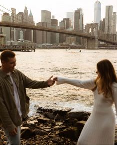 a man and woman holding hands near the water in front of a large cityscape