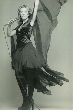 a woman in black dress holding a microphone and wearing high heeled boots with long flowing fabric