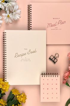 the recipe book is next to flowers and a notepad on a pink surface with writing