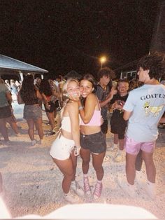 two young women hugging each other on the beach at night with people in the background