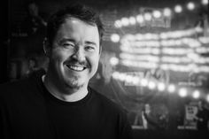 a black and white photo of a man smiling at the camera with lights in the background