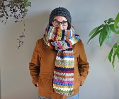 a woman wearing glasses and a multicolored crocheted scarf stands in front of a potted plant