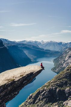 a person sitting on the edge of a cliff overlooking a body of water with mountains in the background