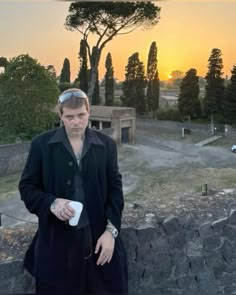 a man standing next to a stone wall with trees in the background and a sunset behind him