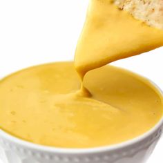 a piece of bread being dipped into a bowl of cheese sauce with a spoon in it