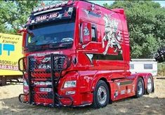 a large red semi truck parked on top of a grass covered field with trees in the background