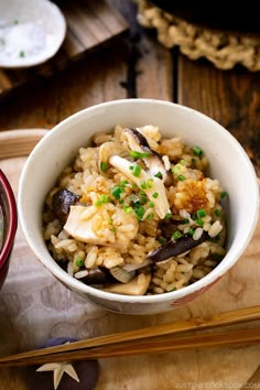 a white bowl filled with rice and mushrooms on top of a wooden table next to chopsticks