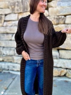 a woman standing in front of a stone wall wearing a brown sweater and blue jeans