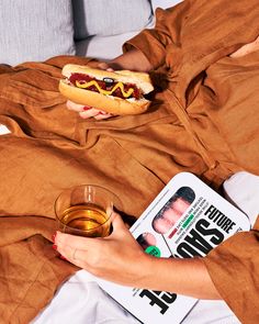 a person laying on a bed with a hot dog and a glass of beer in their hand