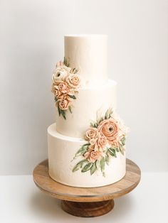 a three tiered wedding cake with flowers on it sitting on a wooden stand in front of a white wall