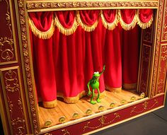 a green frog sitting on top of a wooden floor in front of red velvet curtains