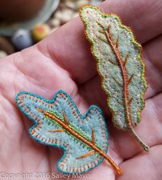 two small embroidered leaves in the palm of someone's hand, one has a green and orange leaf on it