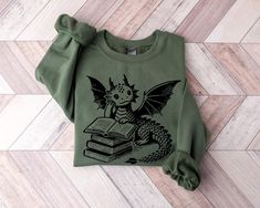 Cozy up with your favorite fantasy novel in our Dragon Book Lover Sweatshirt! This comfy pullover showcases an endearing dragon nestled among books, perfect for chilly days spent reading. Designed for bookworms, fantasy enthusiasts, and dragon fans, it's a warm hug for bibliophiles. Whether you're a librarian, teacher, or just someone who loves to get lost in stories, this sweatshirt is your ideal reading companion. It's not just clothing; it's a statement of your passion for books and imagination. PRODUCT DESCRIPTION: Ideal for any situation, a unisex heavy blend crewneck sweatshirt is pure comfort. These garments are made from polyester and cotton. This combination helps designs come out looking fresh and beautiful. The collar is ribbed knit, so it retains its shape even after washing. T Dragon Sweater, Gifts For Brothers, Dragon Book, Lover Sweatshirt, Sweater Cute, Fantasy Novel, Book Dragon, Bookish Gifts, Warm Hug